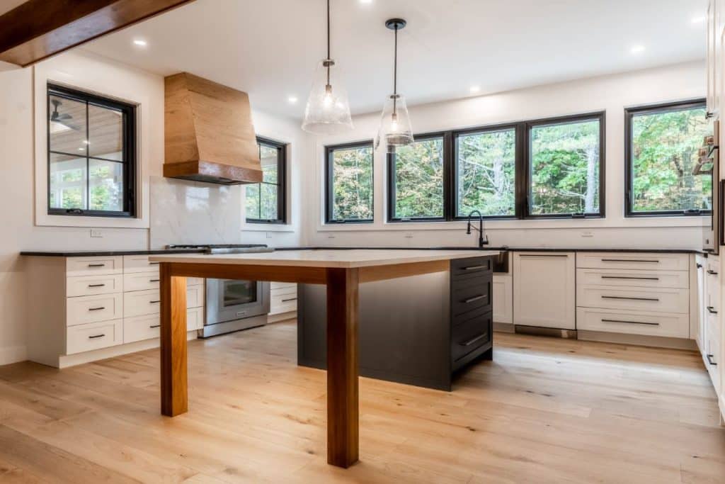 Wide view of a brightly lit kitchen. The sink wall features a wall of windows above a black countertop with a matching black farmhouse sink. The cabinets below are white with black handles. There is another wall of white cabinets, including a wall oven and a paneled white fridge that blends into the cabinets. In the center of the room is the kitchen island, which is open at one end, with walnut-finished legs. The other end of the island is supported a black cabinet with three drawer bases. The island is topped with white quartz and glass pendant lights hang above it. The kitchen is otherwise lit by recessed lighting in the ceiling. The floor below is a light maple with visible knots and wood grain.